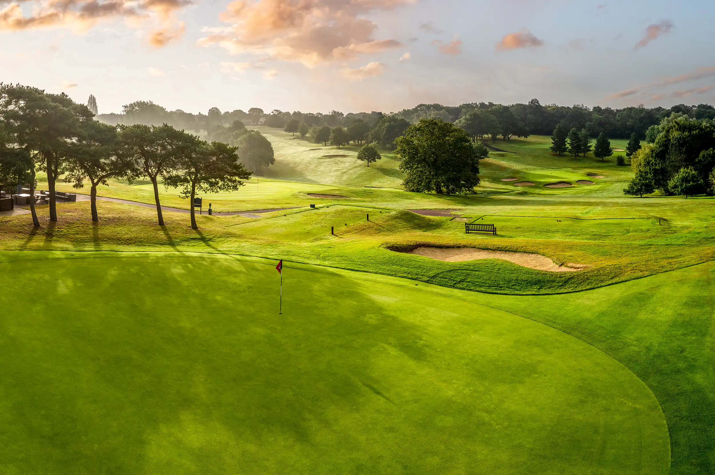 A parkland golf course at sunrise: the green, the flag and fairway stretching into the trees.